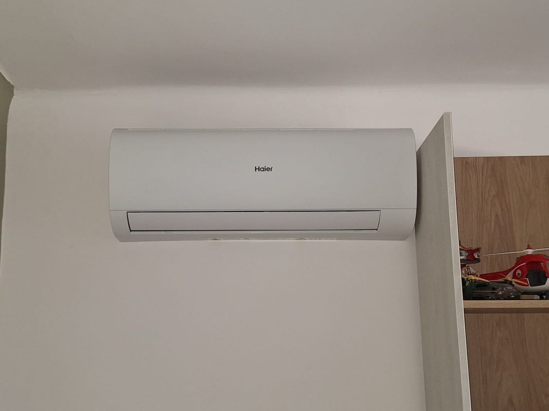 Biały klimatyzator Haier zamontowany na ścianie w pokoju, obok półki z zabawkami, w tym czerwonym helikopterem.