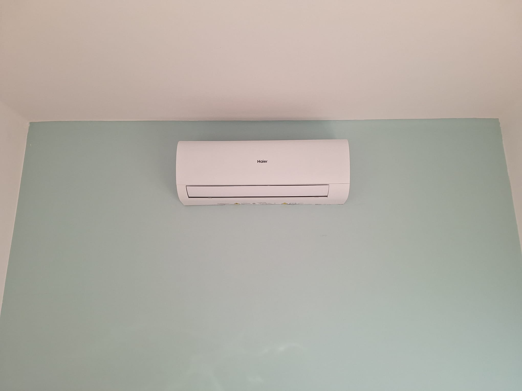 Biała jednostka klimatyzacyjna Haier zamontowana na pastelowo-zielonej ścianie w pomieszczeniu.