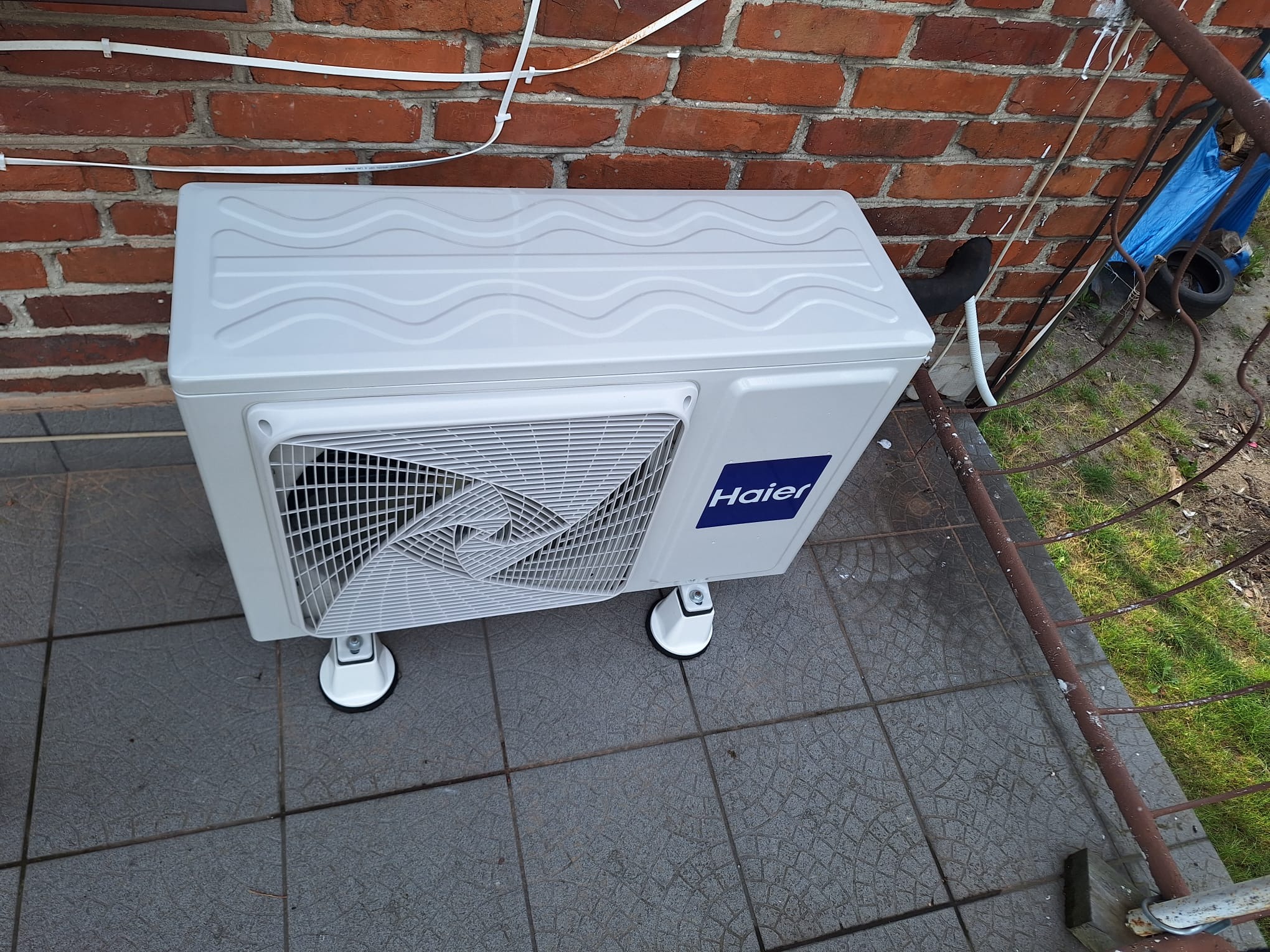 Biała zewnętrzna jednostka klimatyzacji Haier na szarych płytkach tarasowych, z widoczną ceglaną ścianą w tle i fragmentem zardzewiałej barierki.