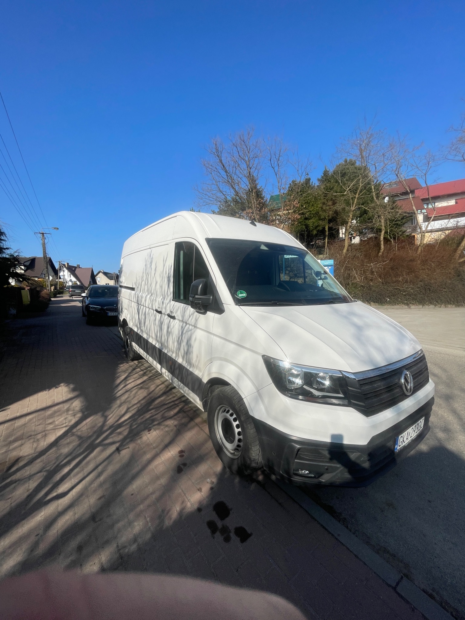 Biały samochód dostawczy Volkswagen Crafter zaparkowany na brukowanej ulicy w słoneczny dzień, z widocznymi cieniami drzew na chodniku i innymi pojazdami w tle.