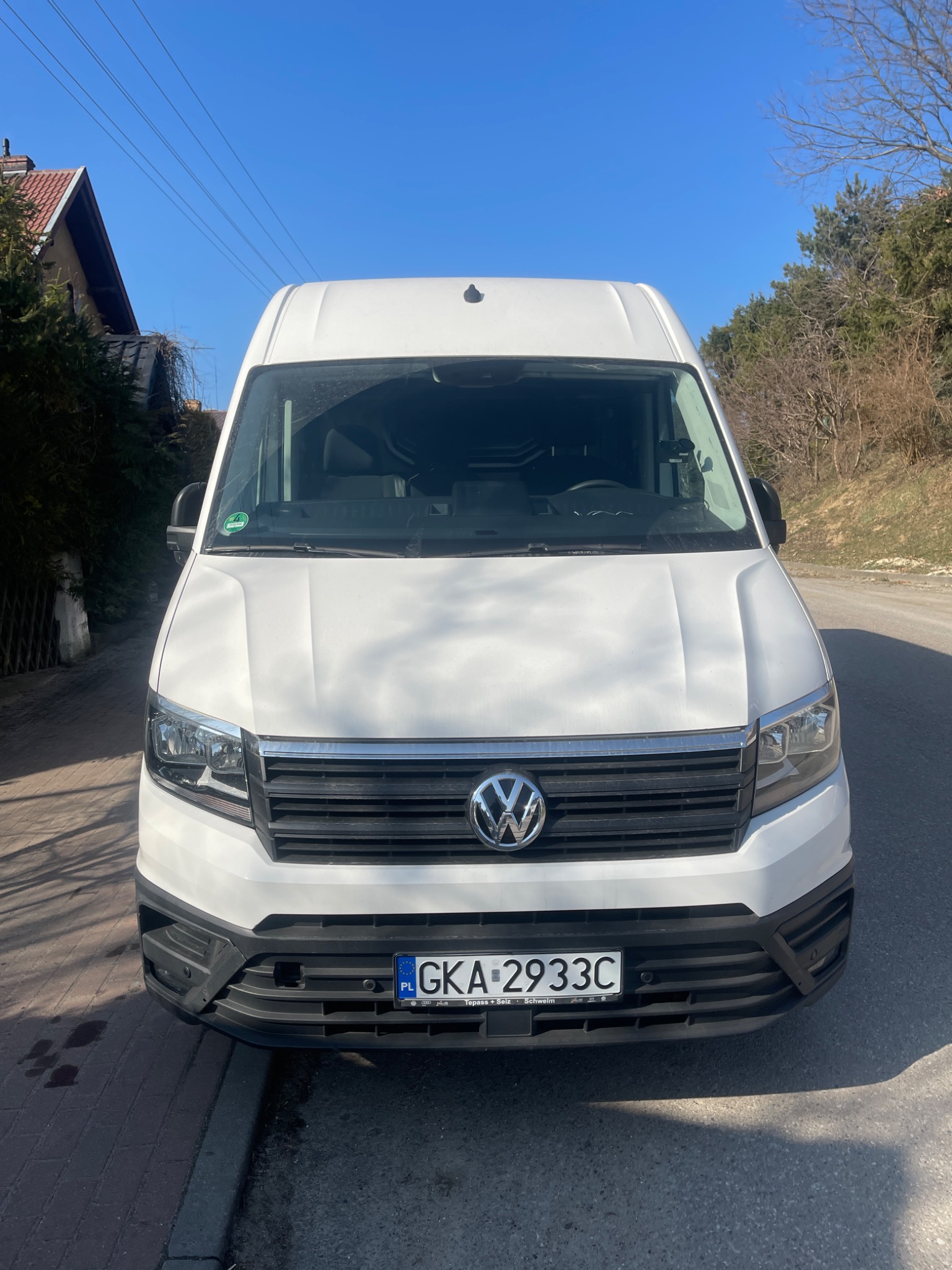 Biały Volkswagen Crafter zaparkowany na asfalcie, z widoczną tablicą rejestracyjną GKA 2933C i zieloną naklejką ekologiczną na szybie, sfotografowany w słoneczny dzień z perspektywy przodu pojazdu.