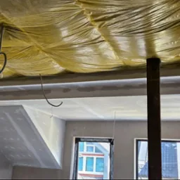 Poddasze w trakcie ocieplania od wewnątrz, widoczna żółta folia izolacyjna pod dachem, elementy konstrukcyjne i ściany w stanie surowym, przygotowane do dalszych prac wykończeniowych.