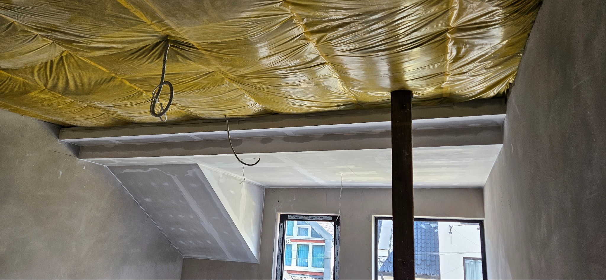 Poddasze w trakcie ocieplania od wewnątrz, widoczna żółta folia izolacyjna pod dachem, elementy konstrukcyjne i ściany w stanie surowym, przygotowane do dalszych prac wykończeniowych.