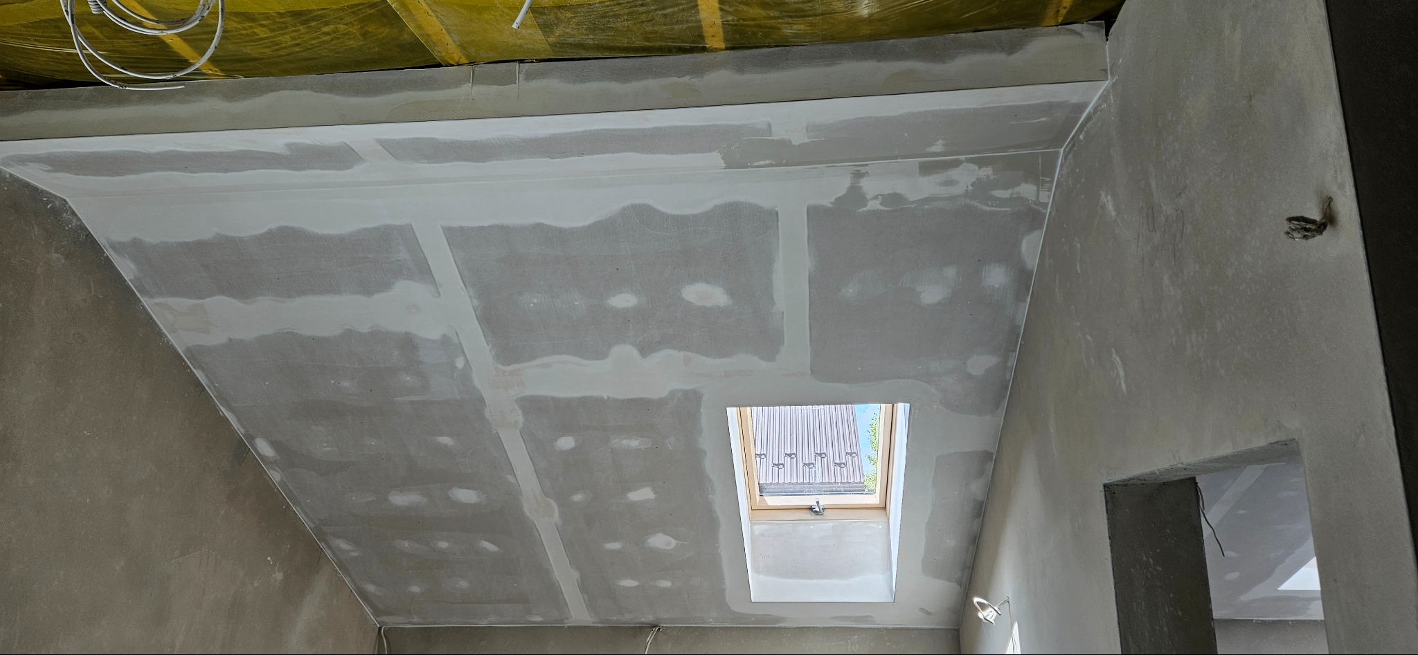 Poddasze z oknem dachowym w trakcie prac wykończeniowych; widoczne łączenia płyt gipsowo-kartonowych wzmocnione taśmą i częściowo pokryte szpachlą, surowe ściany.