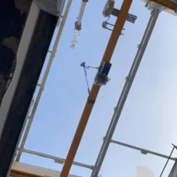 Widok z dołu na konstrukcję stalową budynku w trakcie budowy, z widocznym żurawiem wieżowym i podwieszonym urządzeniem dźwigowym, wszystko na tle błękitnego nieba.