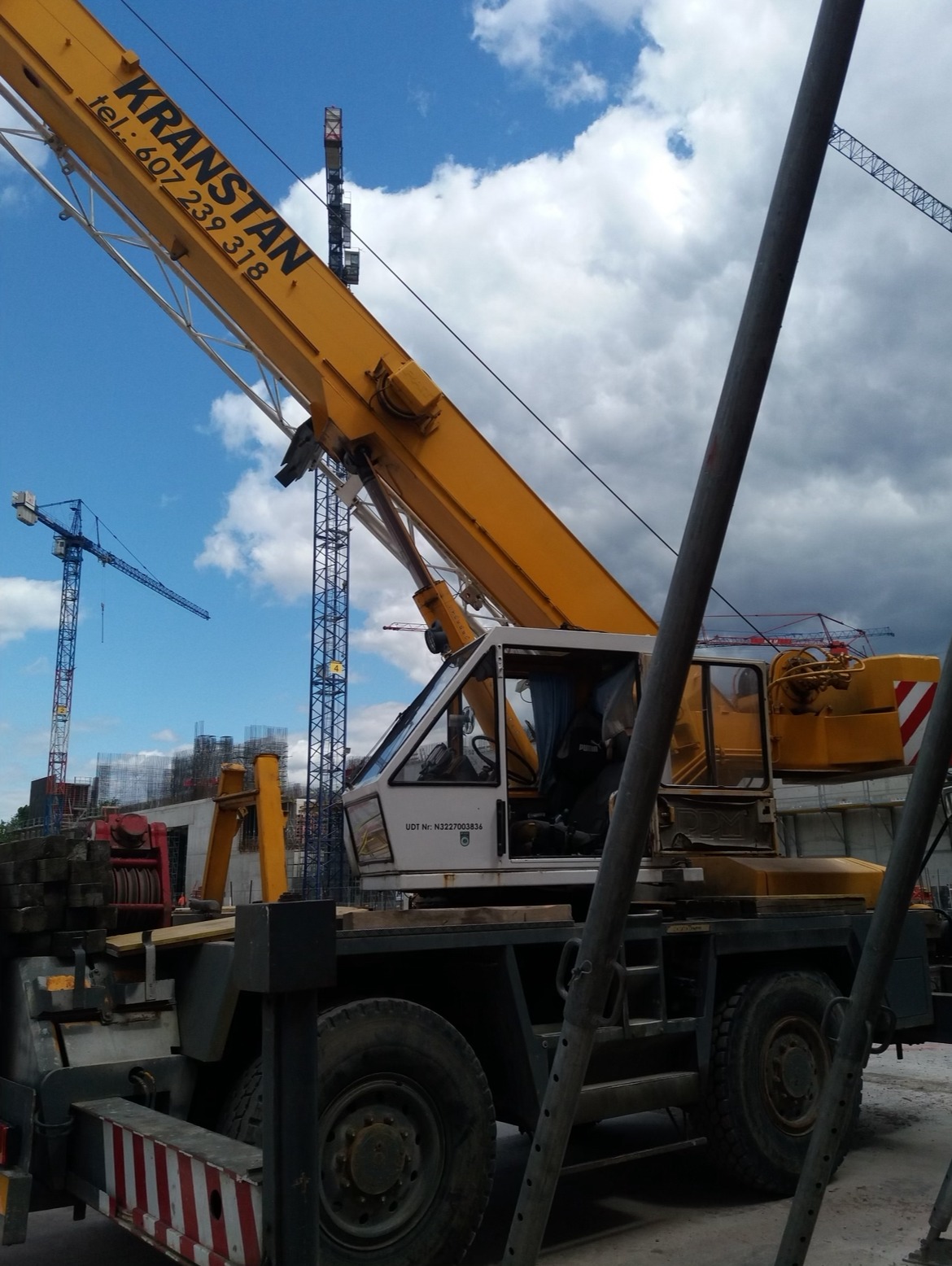 Żółty dźwig samochodowy marki KRANSTAN na placu budowy, z widocznymi elementami konstrukcyjnymi w tle i innymi dźwigami w oddali, uchwycony pod pochmurnym niebem.