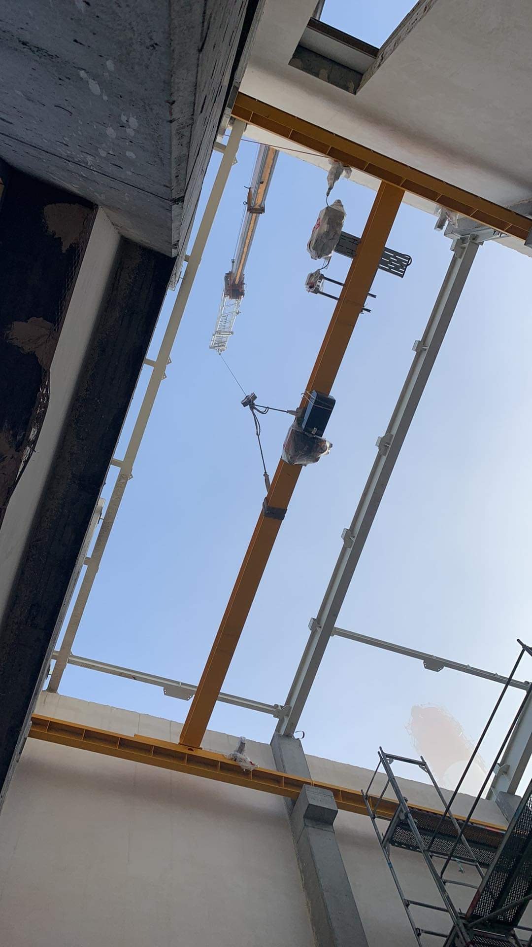 Widok z dołu na konstrukcję stalową budynku w trakcie budowy, z widocznym żurawiem wieżowym i podwieszonym urządzeniem dźwigowym, wszystko na tle błękitnego nieba.