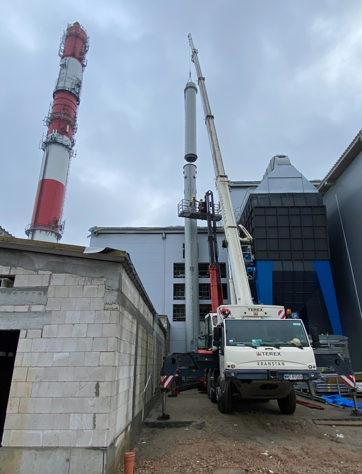 Montaż segmentu komina przemysłowego za pomocą dźwigu Terex Kranstan, pracownicy w koszu na wysokości, w tle budynki przemysłowe i drugi komin w czerwono-białe pasy.