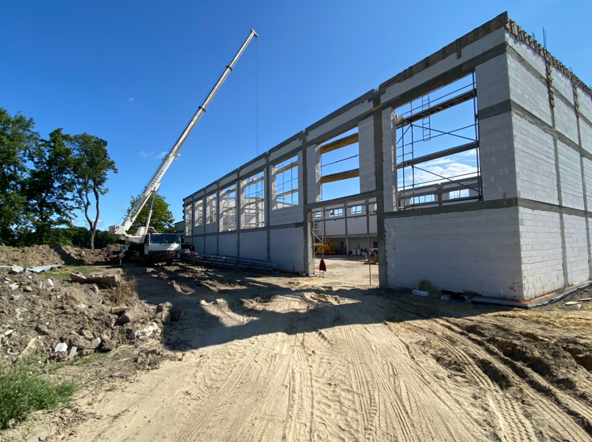 Budowa hali: biały dźwig teleskopowy ustawiony obok wznoszonej konstrukcji z betonu komórkowego, widoczne ślady opon na piaszczystym placu budowy.