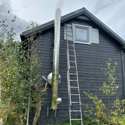 ARKADIUSZ NAWROCKI ZAKŁAD USŁUG INSTALACYJNYCH LECH-AR - Montaż komina zewnętrznego ze stali nierdzewnej przy ciemnym, drewnianym domu. Widoczna drabina i osoba uszczelniająca połączenia. Dym wydobywa się z komina.