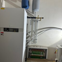 ARKADIUSZ NAWROCKI ZAKŁAD USŁUG INSTALACYJNYCH LECH-AR - Biały piec Viessmann z niebieskim i szarym zbiornikiem wyrównawczym, rury instalacji CO oraz rozdzielacz ogrzewania podłogowego w białej obudowie.