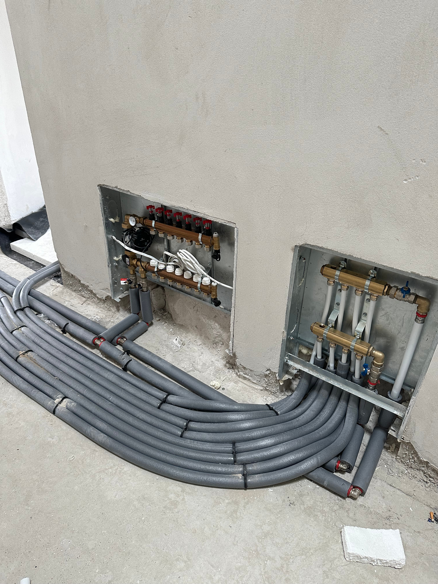 Szare rury ogrzewania podłogowego w izolacji, prowadzone do rozdzielaczy w ścianie. Widoczne elementy instalacji i surowy stan wykończenia.