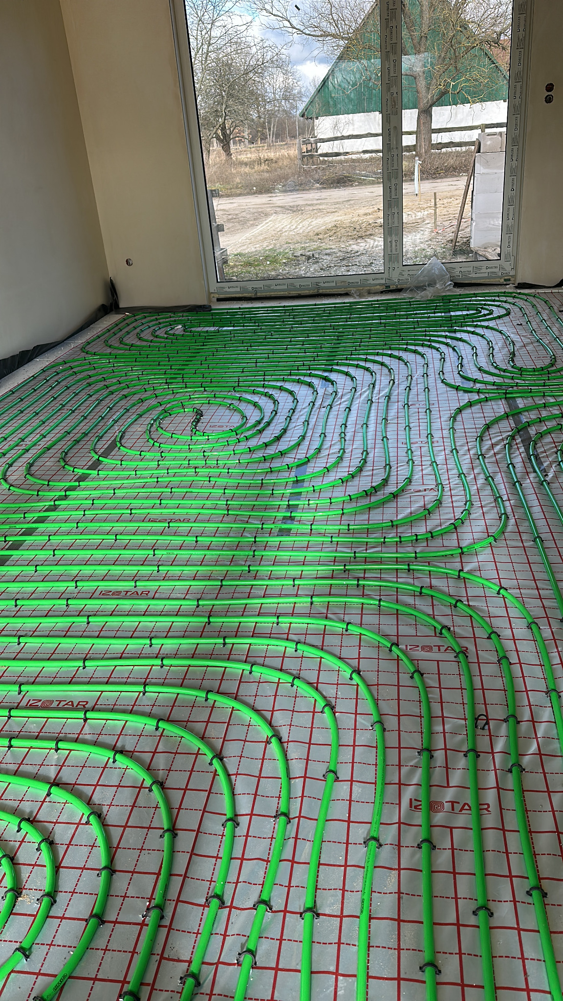 Zielone rury ogrzewania podłogowego ułożone na folii z czerwoną siatką w jasnym pomieszczeniu z dużym oknem. Widoczne mocowania rur.