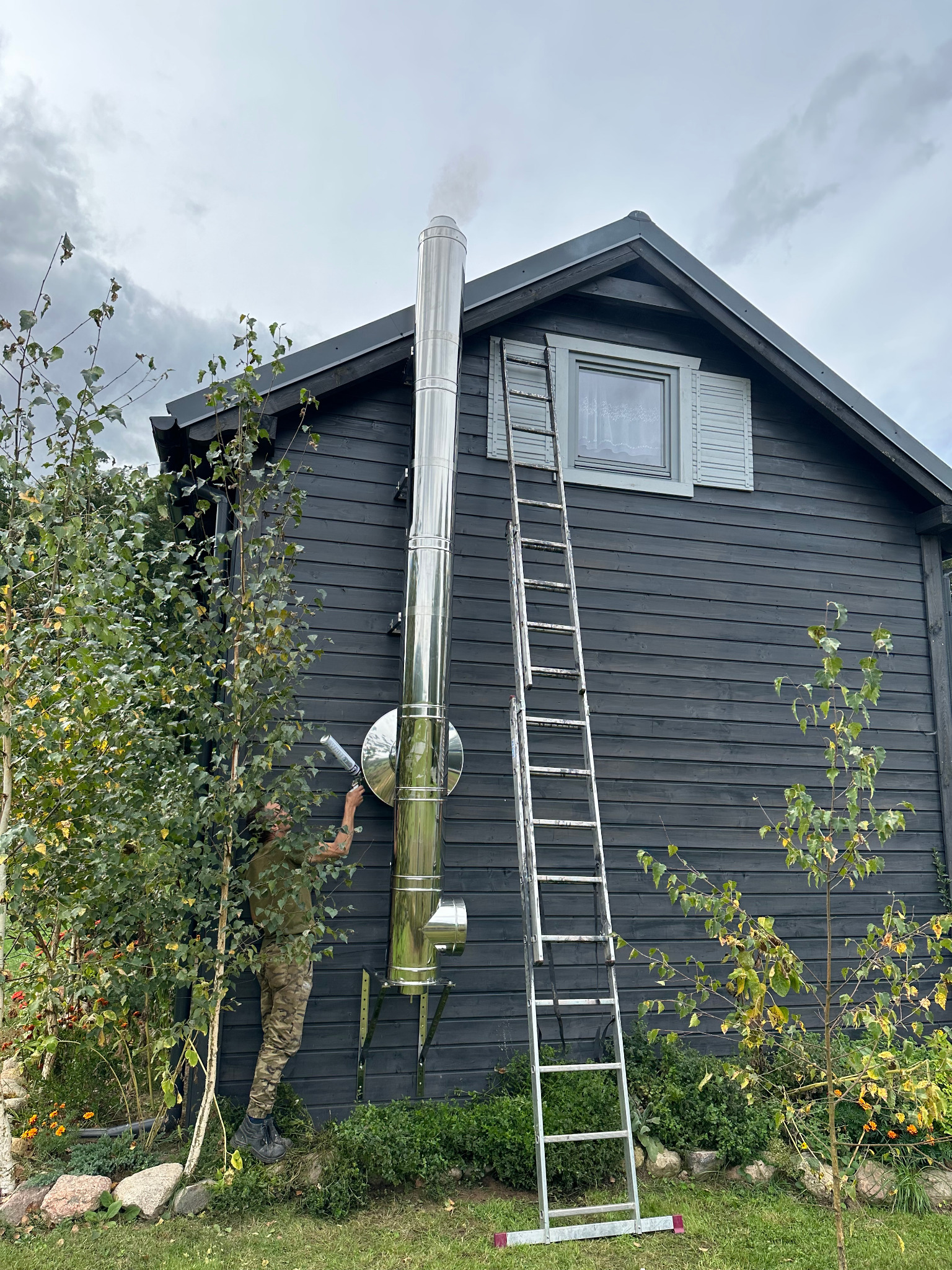 Montaż komina zewnętrznego ze stali nierdzewnej przy ciemnym, drewnianym domu. Widoczna drabina i osoba uszczelniająca połączenia. Dym wydobywa się z komina.