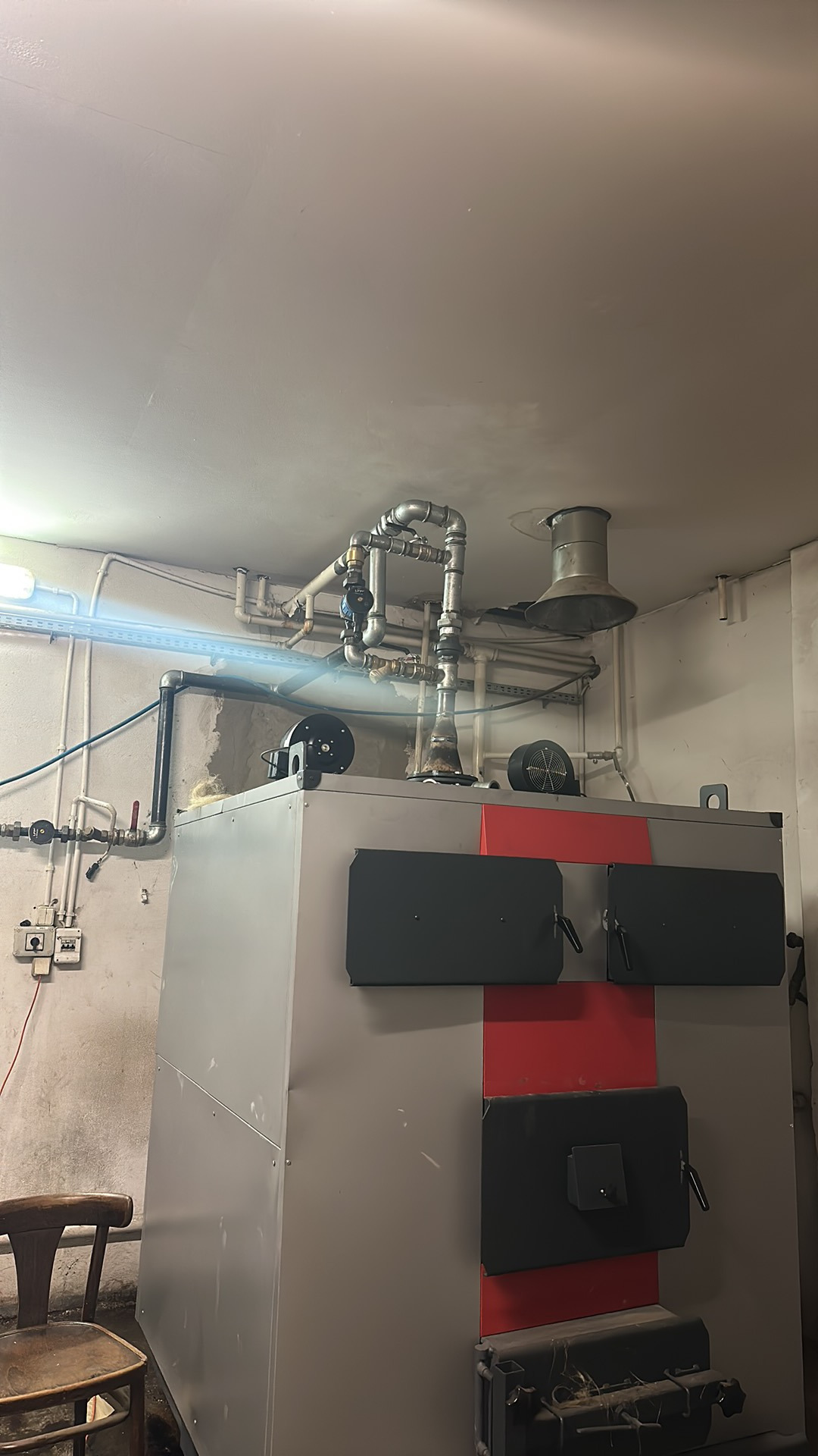 Szary piec CO z czerwonym pasem na tle ściany z instalacją. Widoczne rury, wentylator i fragment krzesła. Ujęcie z dołu, oświetlenie sztuczne.