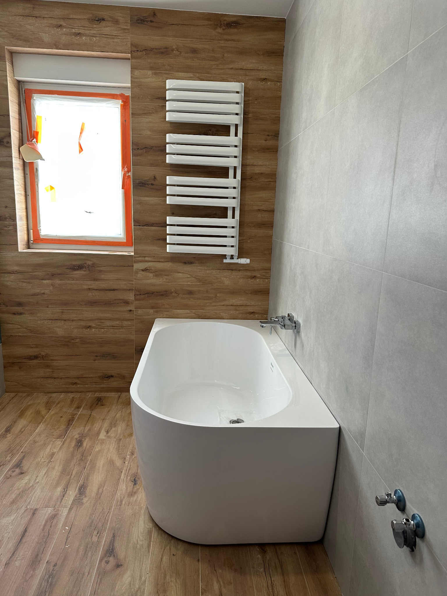 Nowoczesna łazienka z wanną, grzejnikiem drabinkowym i oknem. Płytki imitujące drewno na podłodze i ścianie, szare płytki na sąsiedniej ścianie. Minimalistyczny design.