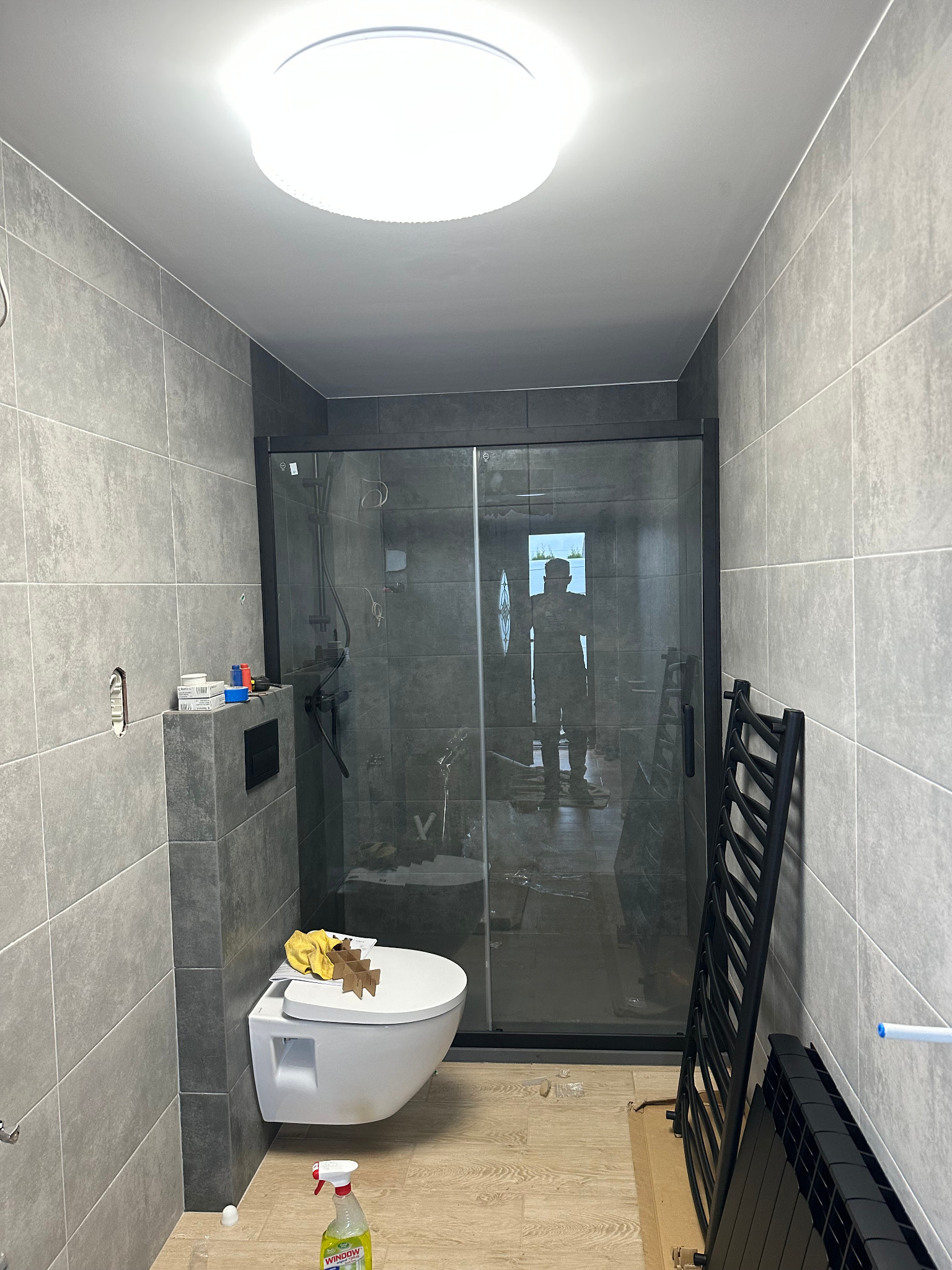 Nowoczesna łazienka w szarościach z kabiną prysznicową typu walk-in, podwieszaną toaletą i grzejnikiem drabinkowym. Widoczna butelka środka czyszczącego.