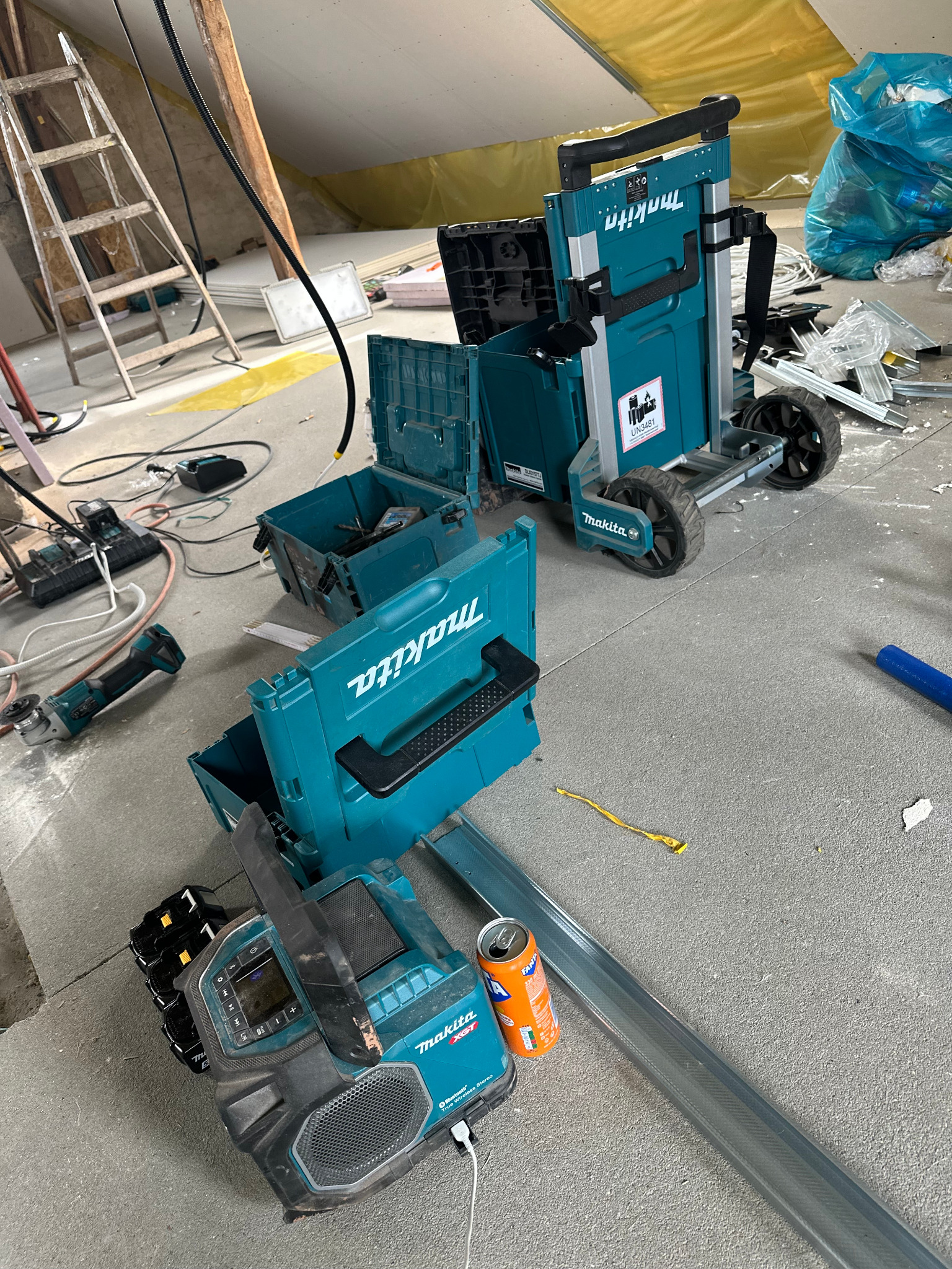 Rozłożone narzędzia Makita na poddaszu w trakcie prac instalacyjnych. Widoczna drabina, skrzynki narzędziowe, radio, puszka napoju i elementy konstrukcyjne.