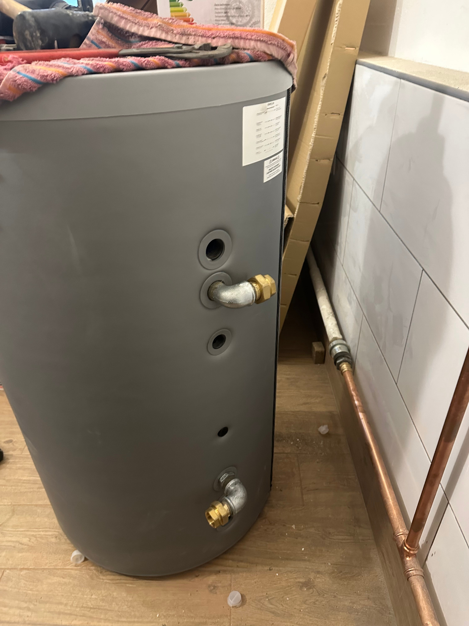 Szary bojler z podłączonymi rurami miedzianymi i elementami instalacji. Widoczne złącza i fragment ściany z białymi płytkami. Ujęcie z bliska, detale instalacji.