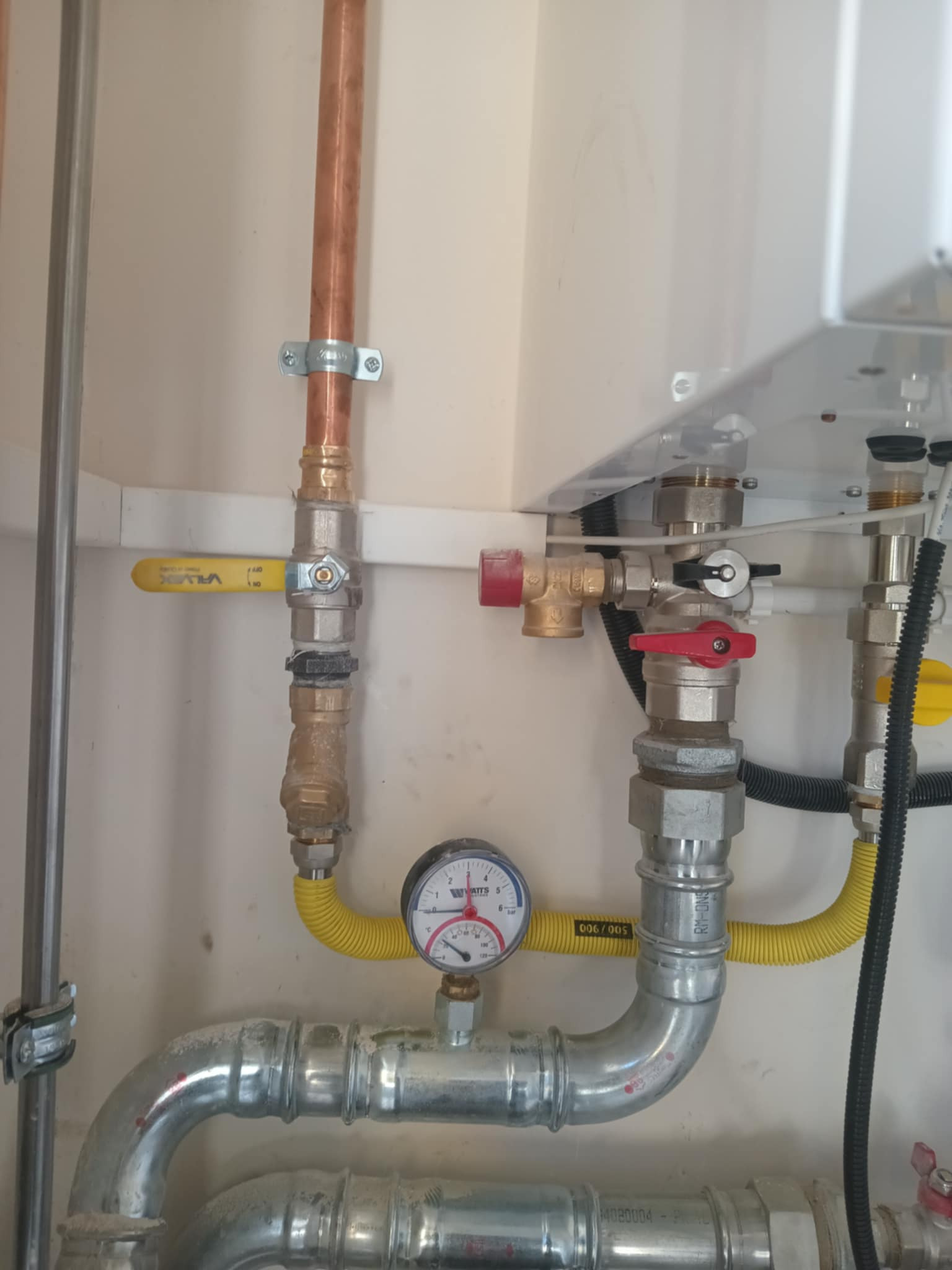 Instalacja gazowa z miedzianymi rurami, zaworami z żółtymi uchwytami i manometrem. Widoczne złącza i elastyczne węże w jasnym pomieszczeniu.