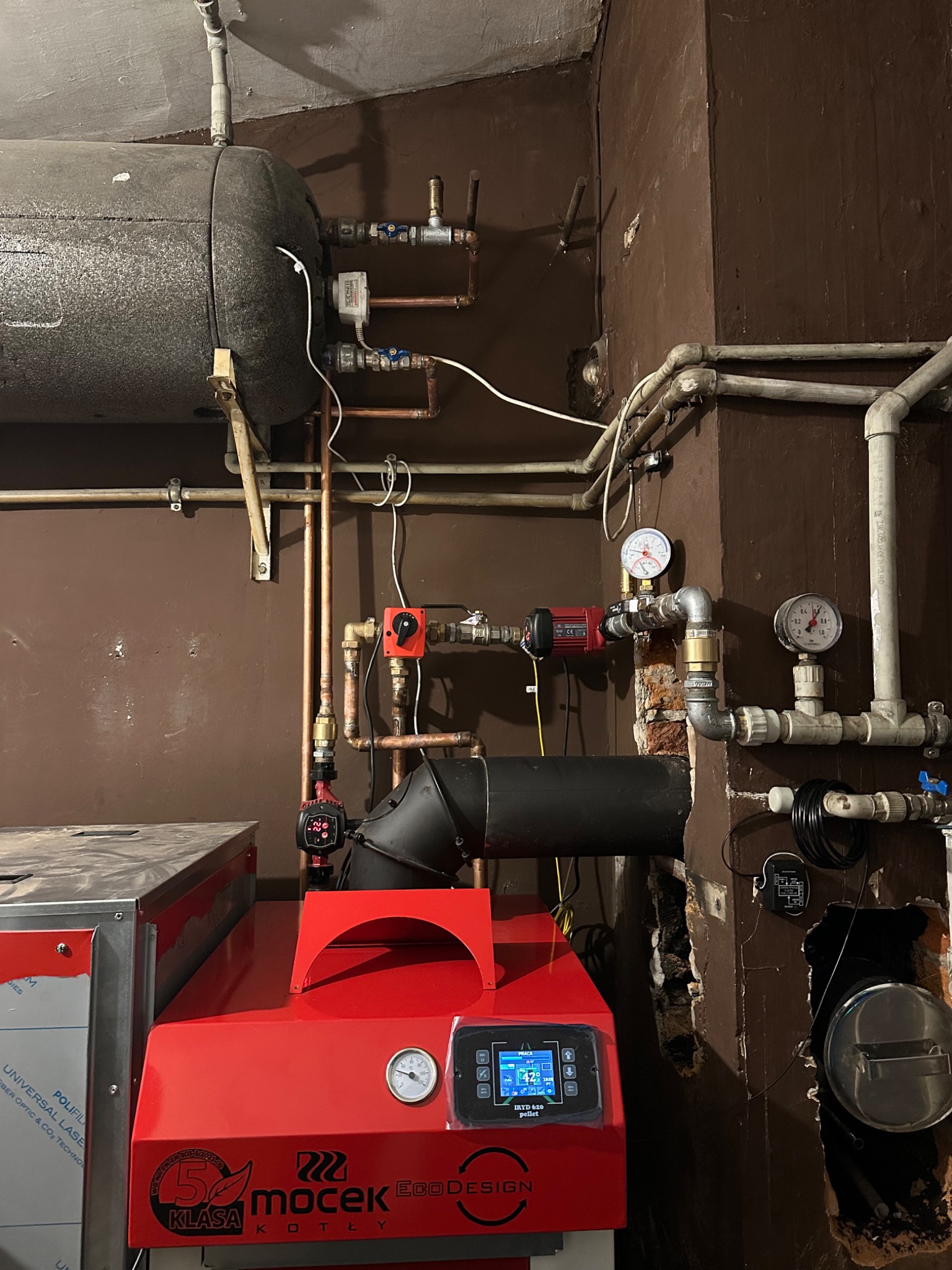 Czerwony piec pelletowy Mocek EcoDesign IRYD 620 z podłączoną instalacją CO w kotłowni o brązowych ścianach. Widoczne rury miedziane i manometry.