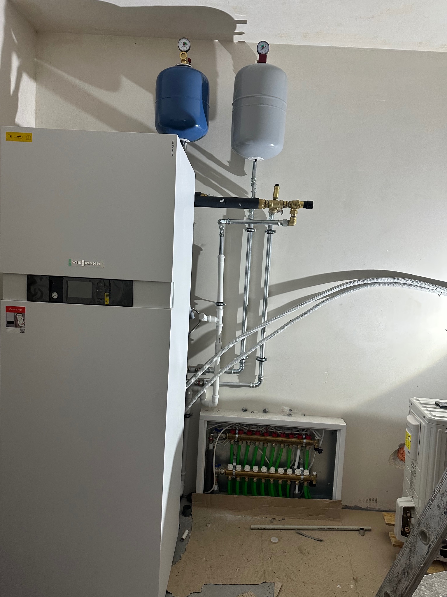 Biały piec Viessmann z niebieskim i szarym zbiornikiem wyrównawczym, rury instalacji CO oraz rozdzielacz ogrzewania podłogowego w białej obudowie.