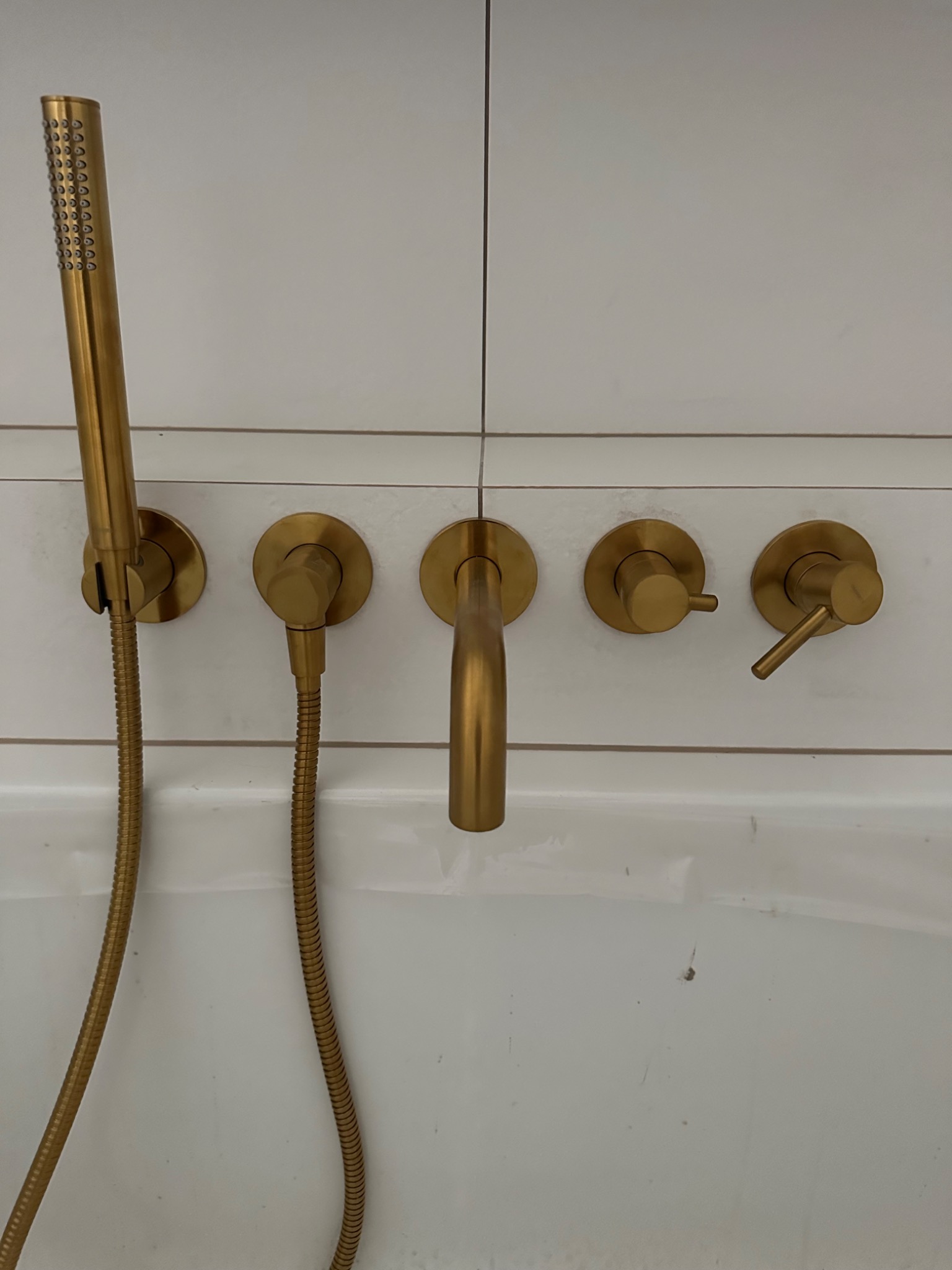 Złota armatura łazienkowa: bateria wannowa z deszczownicą i kurkami, zamontowana na białej, kafelkowanej ścianie nad wanną. Minimalistyczny design, nowoczesne wykończenie.