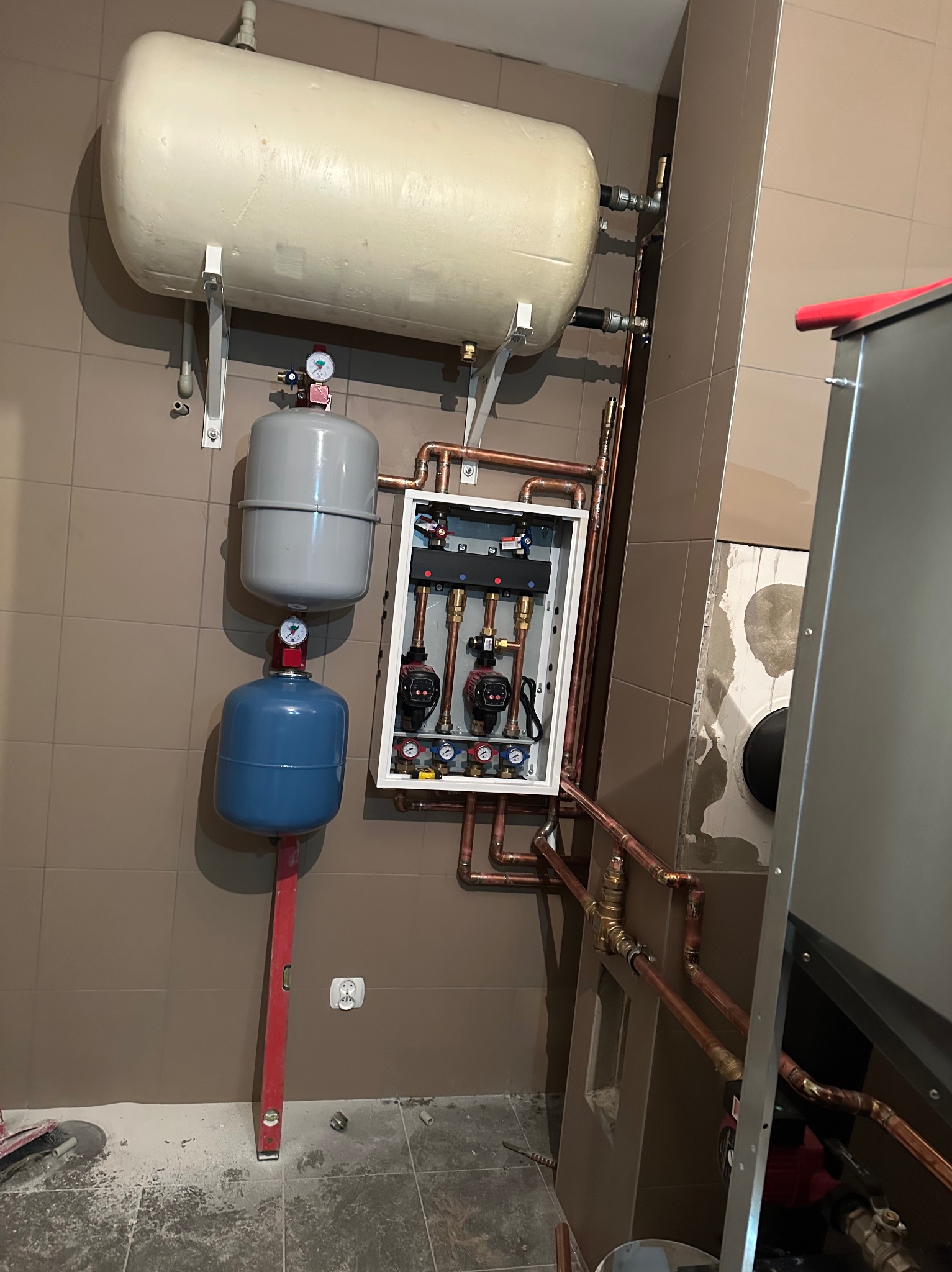 Kompleksowa instalacja CO z zbiornikami wyrównawczymi i rozdzielaczem, rury miedziane, poziomica. Brązowe płytki na ścianie.