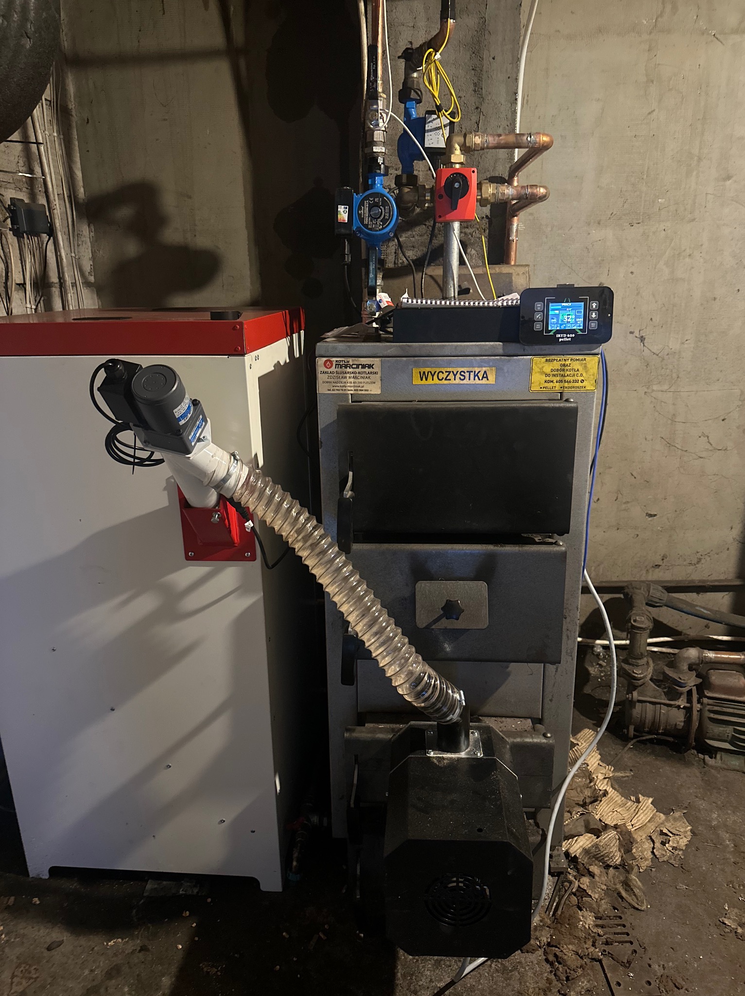 Nowoczesny piec CO na pellet z automatycznym podajnikiem i sterownikiem, zainstalowany w kotłowni. Widoczne rury i armatura instalacji grzewczej.