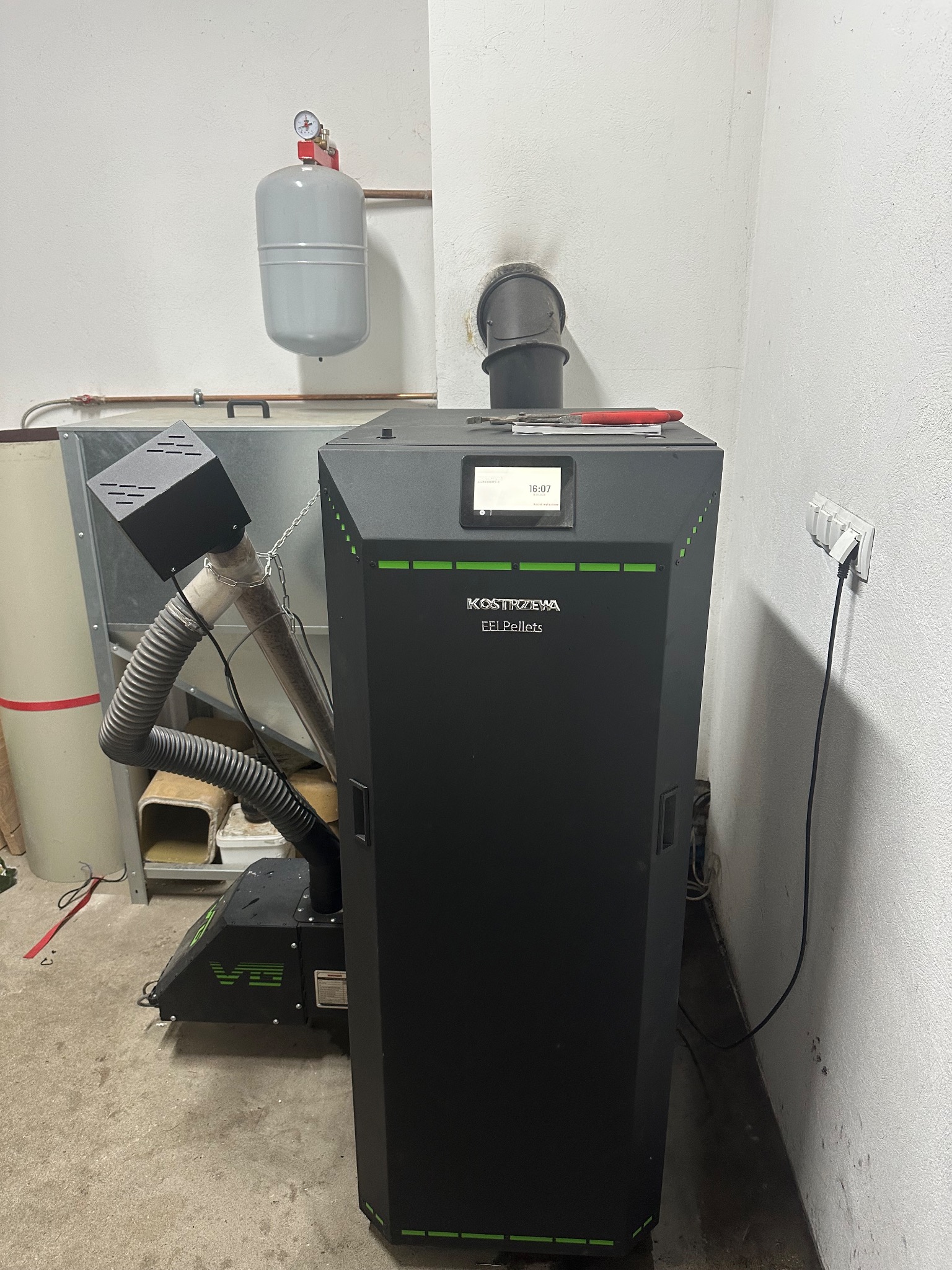 Czarny piec na pellet Kostrzewa EEI Pellets z podajnikiem i zbiornikiem, zainstalowany w kotłowni. Widoczny manometr i rura odprowadzająca spaliny.