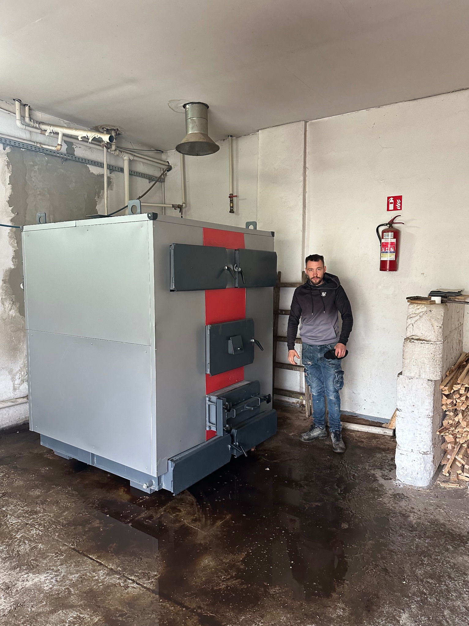 Solidny, nowy piec CO na paliwo stałe, z czerwonymi akcentami, w kotłowni. Obok stoi mężczyzna. Widoczny gaśnica i zapas drewna. Podłoga mokra.
