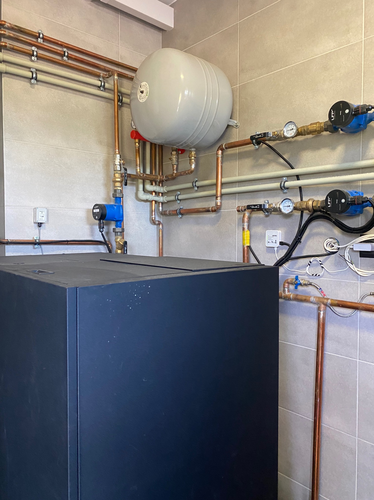 Złożona instalacja grzewcza z kotłem, zbiornikiem wyrównawczym i rurami miedzianymi na tle płytek ceramicznych. Widoczne pompy obiegowe i manometry.