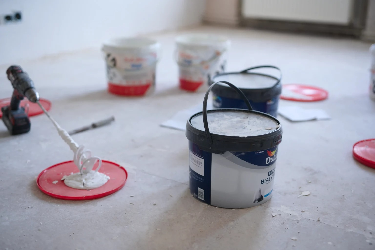 Przygotowanie białej farby Dulux Super Biała do malowania; widoczne wiaderka, mieszadło na wiertarce i podkładki ochronne na podłodze.