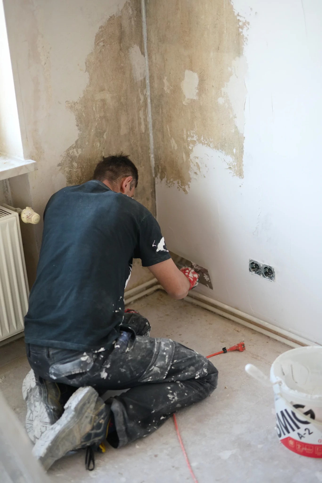 Mężczyzna w ubrudzonych farbą spodniach klęczy i nakłada gładź na ścianę szpachelką, widoczne gniazdka elektryczne i wiadro z farbą.