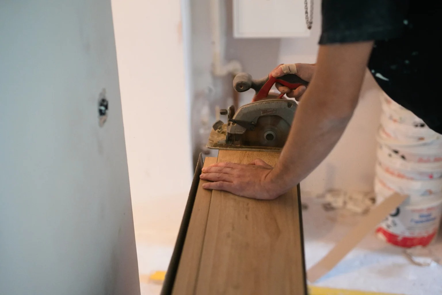 Mężczyzna tnie drewniane panele podłogowe za pomocą elektrycznej piły tarczowej, widoczne białe ściany w trakcie remontu i wiadro z farbą w tle.