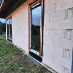 Surowy stan budynku z zamontowanymi oknami z brązowymi ramami i białymi listwami ochronnymi, widoczne bloczki betonowe i fragment dachu.