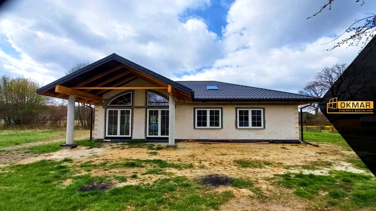 Dom jednorodzinny w trakcie budowy, widoczne białe ramy okienne i przeszklone drzwi tarasowe, ściany z białego bloczka, szara dachówka, logo firmy stolarskiej w prawym górnym rogu.