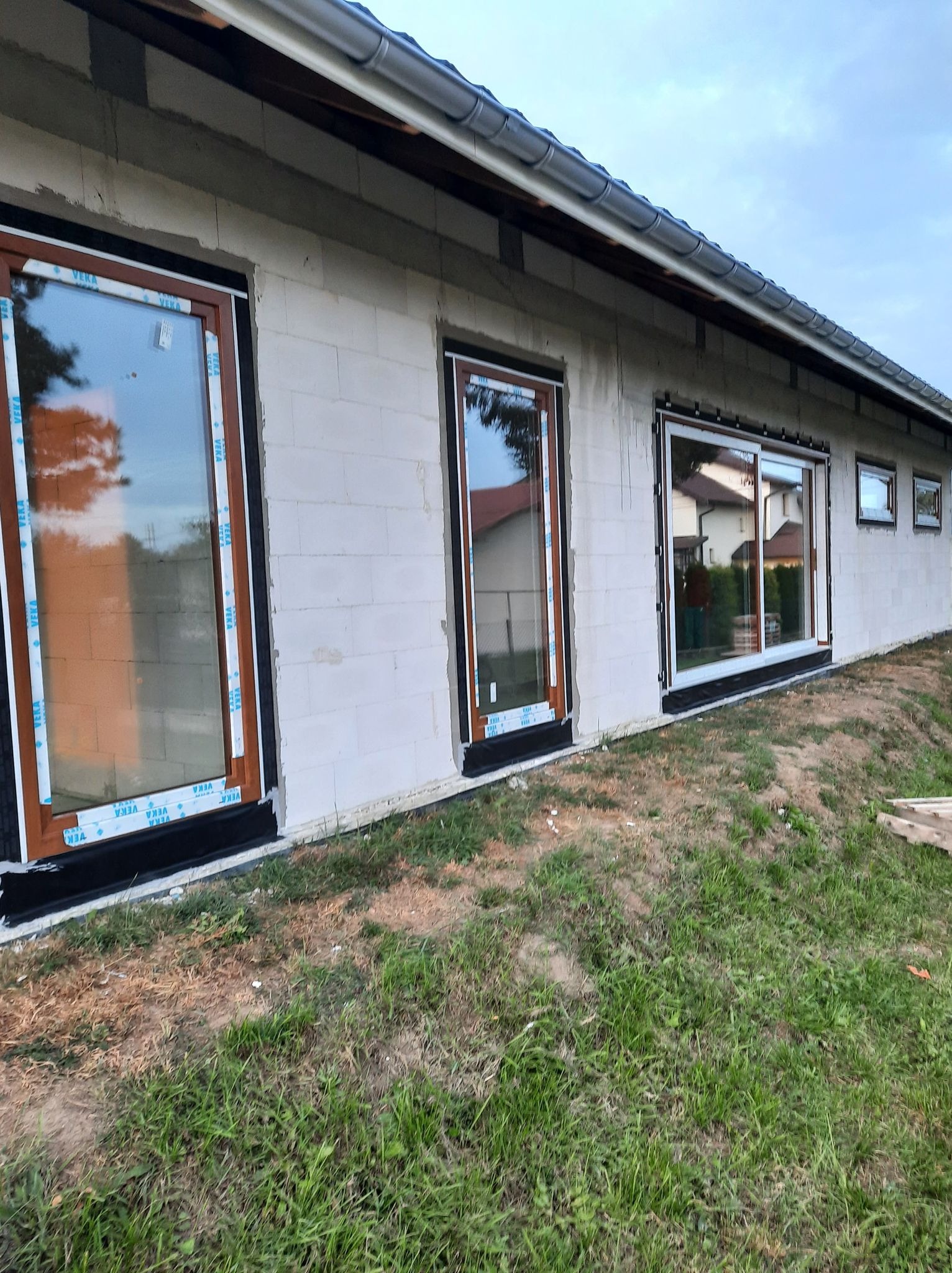 Nowo zamontowane okna i drzwi przesuwne w surowym budynku z betonu komórkowego, widoczne folie ochronne na ramach, teren wokół budynku częściowo porośnięty trawą.
