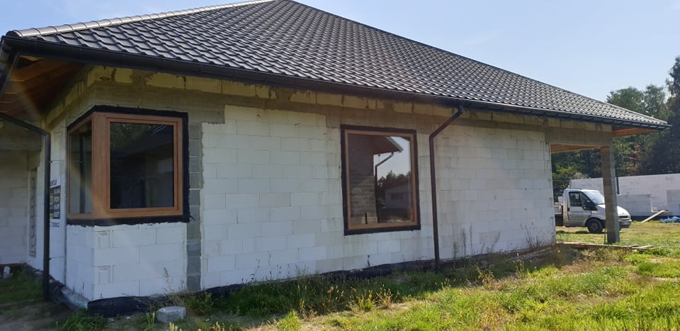 Surowy stan budynku z zamontowanymi oknami PCV o drewnopodobnej ramie, widoczne ocieplenie wokół okien, białe bloczki i ciemna dachówka, na trawniku obok zaparkowany biały dostawczy samochód.