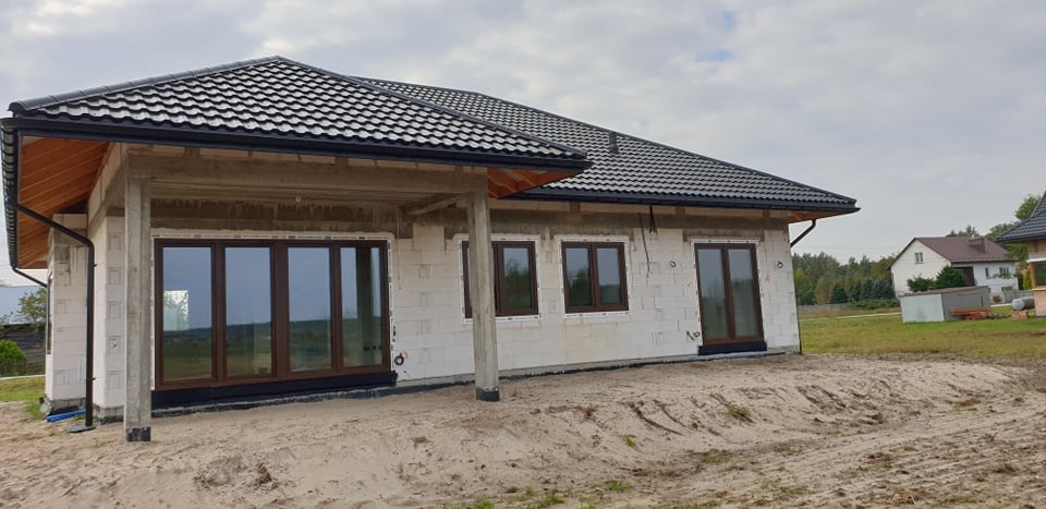 Dom w trakcie budowy z zamontowanymi oknami w brązowych ramach, widoczne bloczki betonowe i czarna dachówka.