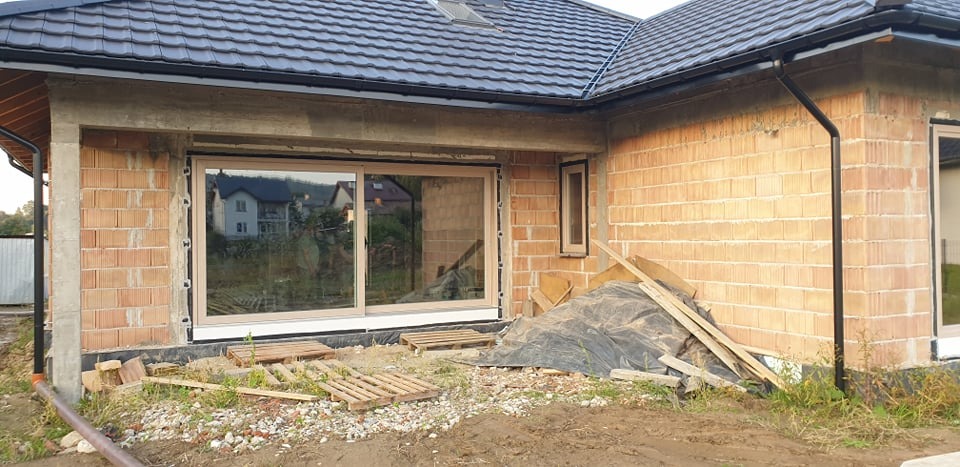 Surowy stan budynku z zamontowanym dużym oknem przesuwnym z jasną ramą, widoczne palety i materiały budowlane na zewnątrz.