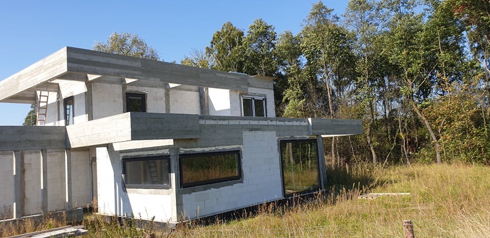 Surowy stan budynku jednorodzinnego z zamontowanymi oknami o czarnych ramach, widok z zewnątrz na tle drzew i trawy.