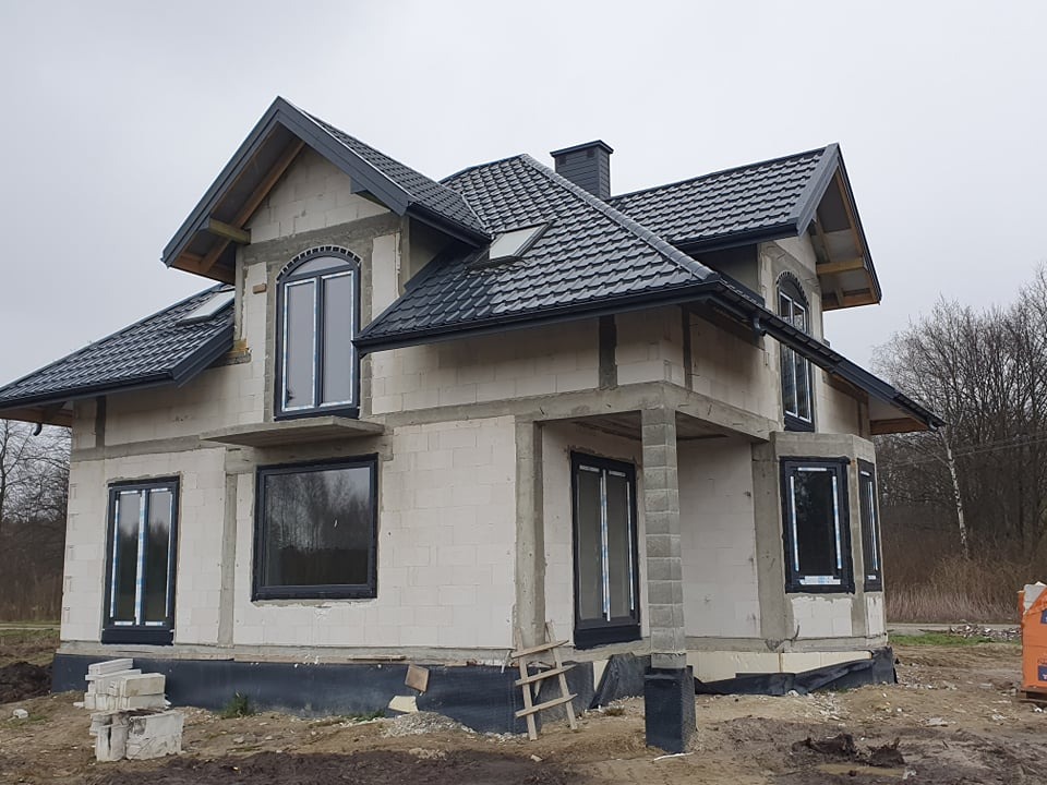 Surowy stan domu w trakcie budowy z zamontowanymi oknami w czarnych ramach, widoczne bloczki betonowe i czarna dachówka, drewniana drabina oparta o ścianę.