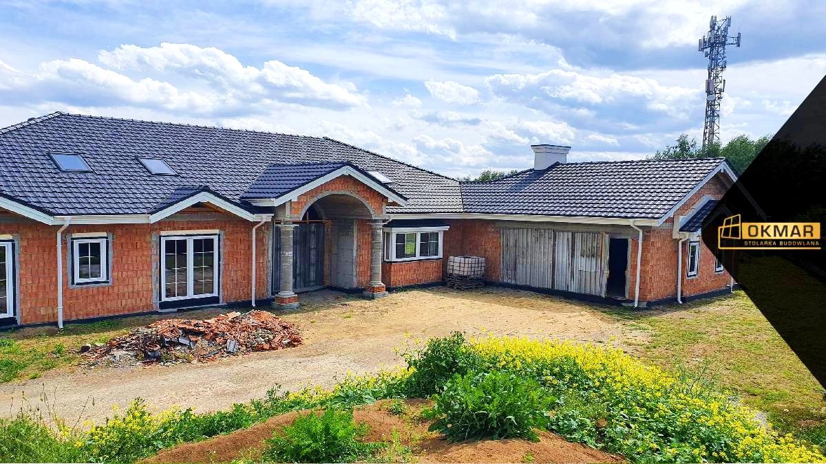 Niedokończony dom jednorodzinny z czerwonej cegły, z białymi oknami i szarą dachówką, widoczne niezabudowane otwory okienne i drzwiowe, na pierwszym planie gruz, w tle maszt telekomunikacyjny.