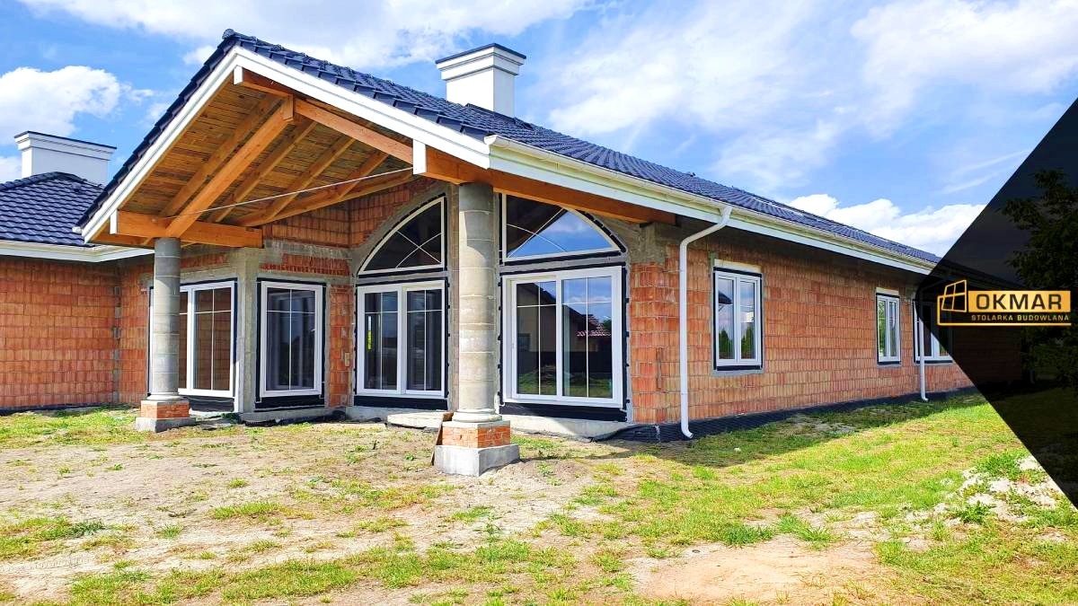 Niedokończony dom z czerwonej cegły z zamontowanymi białymi oknami PCV, widoczne okna łukowe i standardowe, surowy stan wykończenia z odsłoniętą konstrukcją dachu.