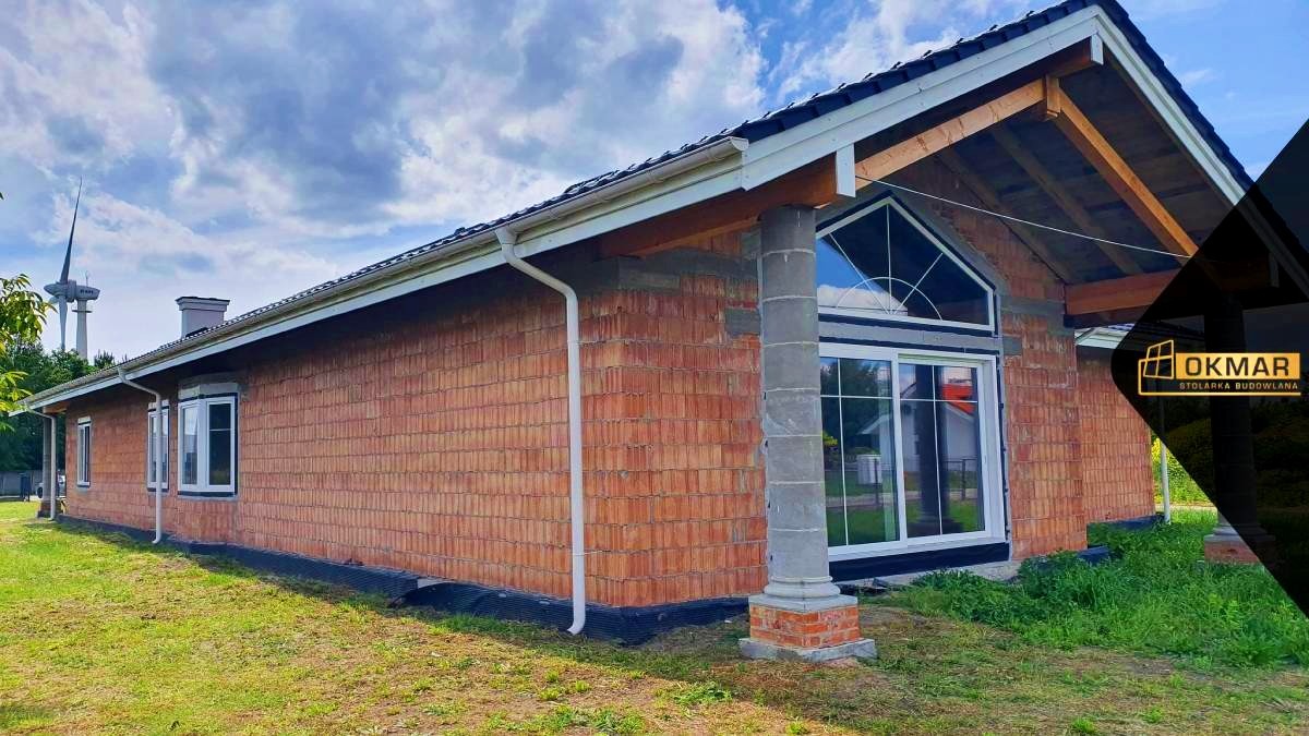 Budynek w trakcie budowy z widocznymi oknami i kolumną z cegły, w tle turbina wiatrowa.