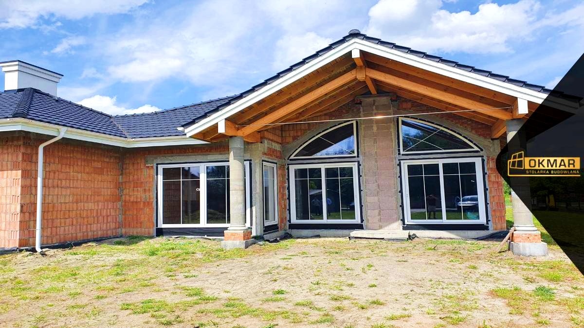 Budynek w trakcie budowy z zamontowanymi oknami o białych ramach, widoczne surowe ściany z cegły i drewniana konstrukcja dachu, na pierwszym planie niezagospodarowany teren.