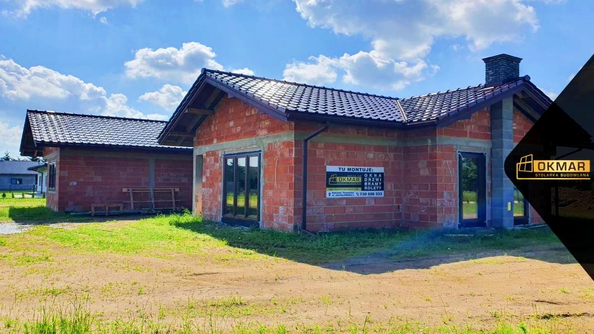 Surowy budynek z czerwonej cegły z zamontowanymi czarnymi oknami, widoczny szyld firmy stolarskiej OKMAR, błękitne niebo z chmurami w tle.