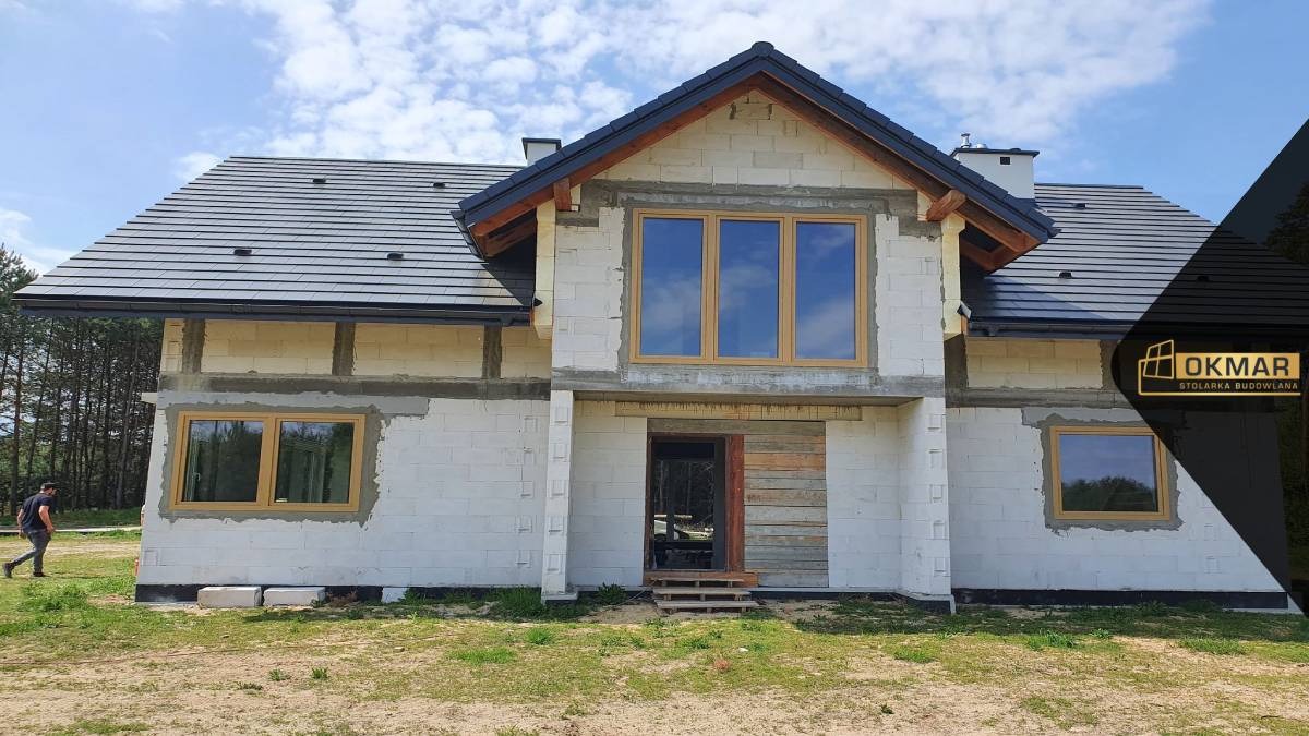 Surowy stan domu jednorodzinnego z zamontowanymi oknami o drewnianej ramie, widoczny mężczyzna w oddali oraz logo firmy w prawym górnym rogu.