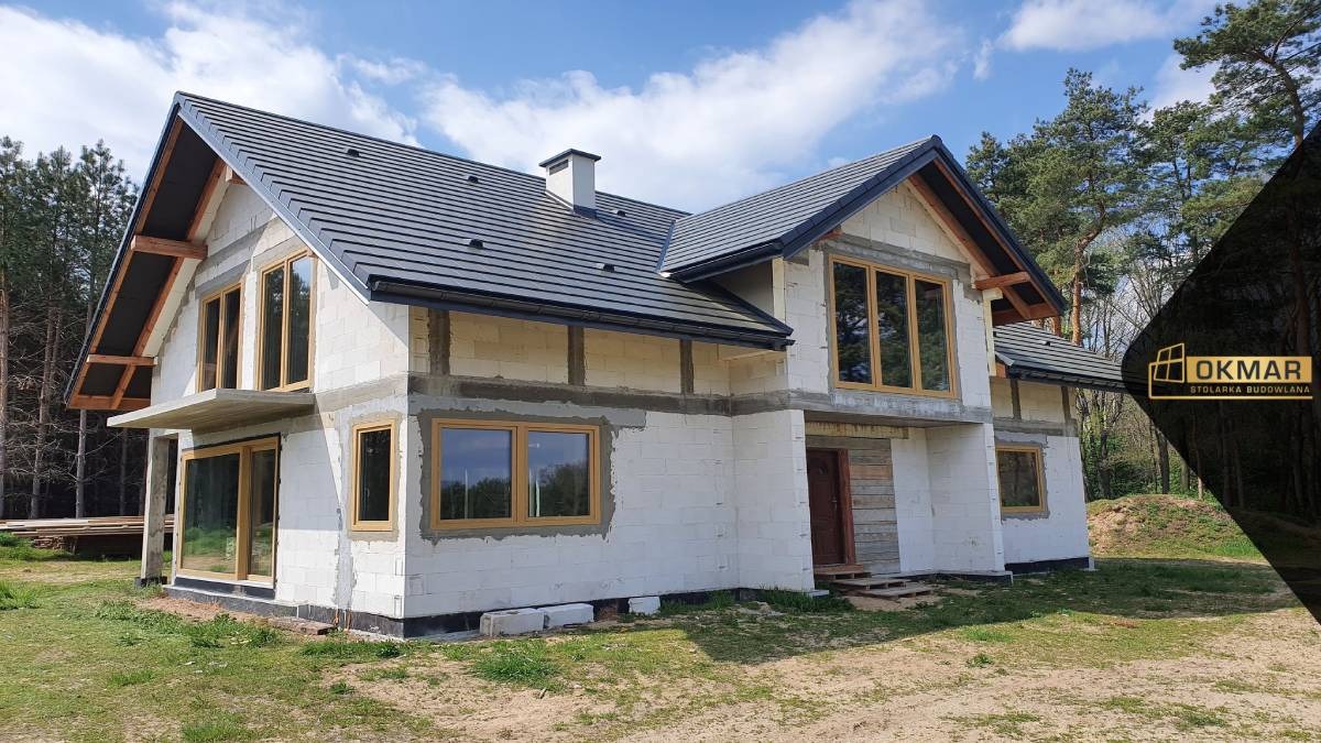 Surowy stan budynku jednorodzinnego z zamontowanymi oknami w drewnianej ramie, widoczne białe bloczki, szara dachówka i las w tle.